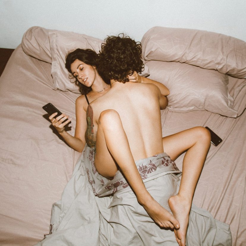 Men on top of a woman in bed while she is on her phone